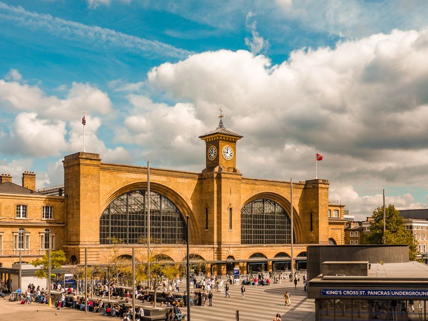 A large historic train station building made of golden-brown brick with a prominent clock tower featuring a pointed spire stands against a partly cloudy sky with white and gray clouds. The front facad