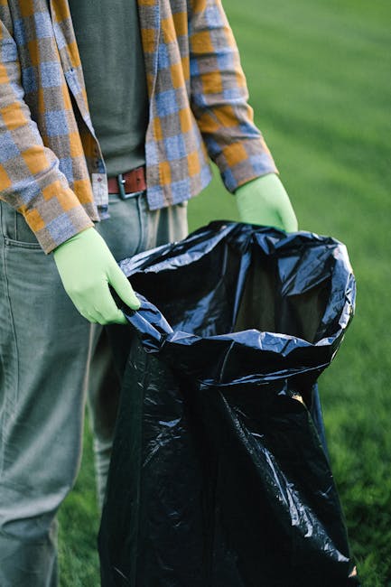 A person wearing a plaid shirt with yellow, gray, and blue tones, along with gray trousers and light green gloves, is holding open the top of a large black rubbish bag made of plastic material, which 